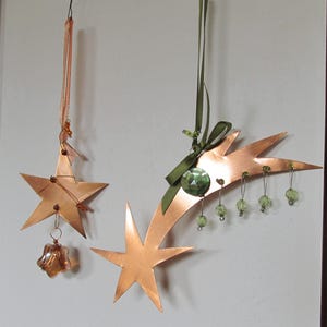 Op de afbeelding: Drie koperkleurige sterornamenten hangen tegen een neutrale muur. Eén is een vijfpuntige ster met een bungelend glazen ornament. Een ander is een vallende ster vorm met groene kralen. De derde is een vijfpuntige ster.