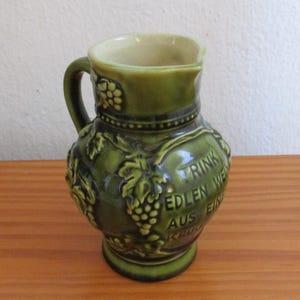 Puede incluir: Una jarra de cerámica verde con un diseño en relieve de racimos de uvas y hojas. El cuerpo de la jarra muestra el texto en relieve TRINK EDLEN WEIN AUS EINEM KRUG. Tiene un asa curva y un pico vertedor ancho y abierto con interior crema.