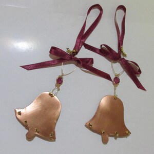 Könnte beinhalten: Zwei kupferfarbene Glockenornamente mit bordeauxfarbenen Schleifen und goldenen Akzenten. Jede Glocke hat einen kleinen, runden, roten Edelstein und ist mit kleinen goldenen Nieten verziert. Die Glocken sind an dünnen Golddrähten aufgehängt.