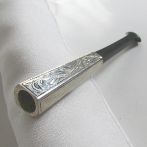 May include: A cigarette holder featuring a hexagonal silver-toned metal section with intricate engraved scrollwork patterns and a smooth, tapered black mouthpiece. The metal portion displays a polished finish with detailed etched lines.