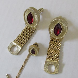 May include: A vintage gold-tone jewelry set featuring a pair of cufflinks and a tie tack. Each piece has a textured, circular design with a red, oval-shaped stone in the center. The cufflinks have a mesh chain design, and the tie tack has a chain.
