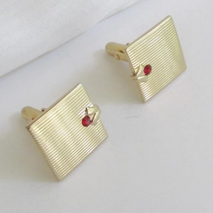 May include: A pair of gold-toned cufflinks with a square, ridged design. Each cufflink features a small, red gemstone accent. The cufflinks are set against a white background.