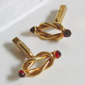 May include: A pair of gold-toned metal cufflinks featuring a twisted knot design. Each cufflink is accented with two small, round, deep red cabochon stones set at the ends of the knot. The cufflinks include a standard hinged toggle closure mechanism.