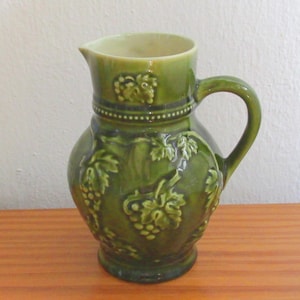 Puede incluir: Una jarra de cerámica de color verde oliva brillante con un patrón en relieve de racimos de uvas y hojas de vid. El recipiente cuenta con un pico ancho, un asa curva y una banda decorativa de cuentas que rodea el cuello, con interior crema.