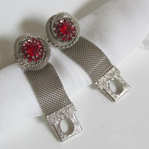 Peut inclure: Une paire de ronds de serviette en métal argenté avec une bande en maille flexible et un sommet décoratif circulaire. Chaque sommet est orné d'une pierre rouge facettée vibrante entourée d'une bordure en métal torsadé et d'une languette plate.