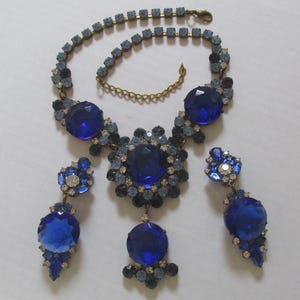 Puede incluir: Un conjunto de collar y pendientes de pedrería azul. El collar presenta un gran colgante de pedrería azul rodeado de pedrería más pequeña. Los pendientes son pendientes colgantes con un gran cristal azul en la parte inferior.