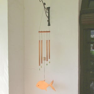 Peut inclure: Un carillon éolien suspendu avec un ornement en forme de poisson. Le carillon comporte des tubes de couleur cuivre suspendus à une barre en bois. Le poisson orange a une queue pointillée et le carillon est suspendu à un crochet en métal décoratif.