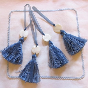 Puede incluir: Cinco borlas de seda azul con acentos circulares blancos y lazos de cinta azul. Las borlas están dispuestas sobre un fondo blanco con un borde azul claro.