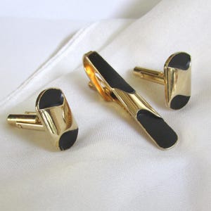 May include: A gold-coloured tie clip and cufflink set with black enamel detailing. The cufflinks and tie clip feature a streamlined, geometric design. The set is presented on a white, textured surface, suggesting a formal or vintage style.