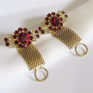 May include: Pair of vintage gold-tone metal napkin rings. Each ring features a central cluster of red rhinestones surrounded by smaller clear and yellow stones. A gold mesh band connects the jewel to a gold ring.