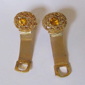 Puede incluir: Un par de pendientes dorados. Cada pendiente presenta un diseño de filigrana con una piedra central de color ámbar. Una banda dorada similar a una malla se extiende hacia abajo, terminando en un lazo rectangular. Los pendientes están diseñados para orejas perforadas.