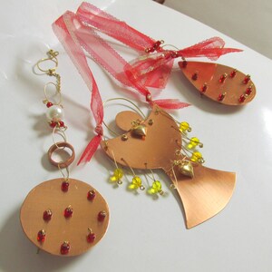 Peut inclure: Trois ornements en métal cuivré comprenant un disque circulaire avec des perles rouges, une forme d'ange avec des perles jaunes et des breloques en forme de cœur doré, et une pièce en forme de goutte avec des perles rouges, suspendus par des rubans rouges.