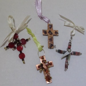 Può includere: Quattro ornamenti a forma di croce fatti a mano. Una croce è fatta di perline rosse, un'altra di rame con filo e pietre, e altre due con rame e perline. Ogni croce ha un nastro o uno spago per appenderla. Le croci sono su sfondo bianco.