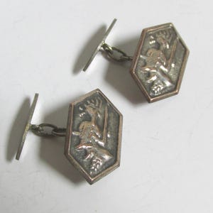 Könnte beinhalten: Ein Paar silberne Manschettenknöpfe im Vintage-Stil. Jeder Manschettenknopf hat ein detailliertes Reliefdesign in einem sechseckigen Rahmen. Das Design scheint eine Figur mit einem Speer und einem Hirsch darzustellen. Die Manschettenknöpfe sind durch eine kleine Kette verbunden.