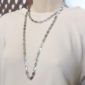 May include: A silver-toned necklace with a double-strand design. The necklace features rectangular, flat metal links. The jewelry is displayed on a cream-colored top, showcasing the necklace's length and style. The necklace is a fashion accessory.