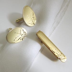 Könnte beinhalten: Ein goldfarbenes Set aus Manschettenknöpfen und einer Krawattenklammer. Die ovalen Manschettenknöpfe und die Krawattenklammer weisen ein eingraviertes Blumendesign auf. Das Set wird auf einem weißen Tuch präsentiert.