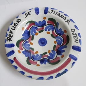 以下が含まれることがあります： 手描きのデザインが施された白いセラミック灰皿。 灰皿には、青、赤、緑、オレンジの花柄と「REFUGIO DE JUANAR OJEN」の文字が描かれています。 リムには青と赤のアクセントがあります。