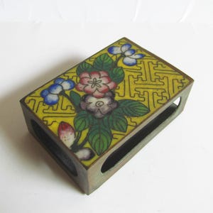 Può includere: Porta fiammiferi rettangolare in ottone con un design floreale giallo. La parte superiore presenta uno sfondo giallo con un motivo ripetuto e fiori colorati. I lati hanno aperture ritagliate.