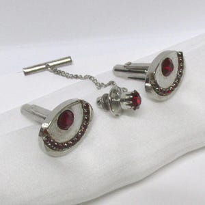 May include: A set of silver cufflinks, a tie tack, and a shirt stud. Each piece has a central oval, deep red stone, encircled by smaller red stones. The set is presented on a white surface.
