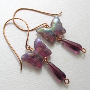 以下が含まれることがあります： 蝶のイヤリングのペア。各イヤリングは、紫と青の色合いの半透明で虹色の蝶のチャームを備えています。蝶の下には、涙の形をした紫色のビーズがぶら下がっています。イヤリングには、銅色のイヤーフックと接続要素が付いています。