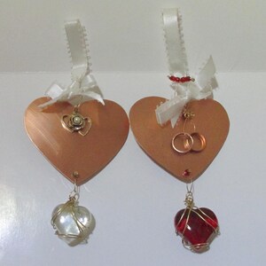 May include: Two copper heart-shaped decorations with white ribbon and bows. One has a gold heart charm with a pearl, the other has two gold rings. One has a white pearl heart, the other a red heart.