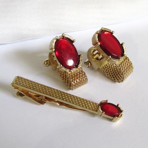 May include: A vintage gold-toned metal jewelry set featuring a tie clip and a pair of cufflinks. Each piece has a large, oval, red gemstone. The cufflinks have a mesh design, and the tie clip has a textured surface.