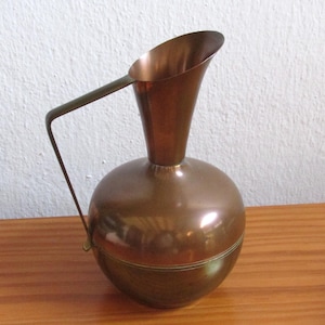 Peut inclure: Une carafe en cuivre poli dotée d'un corps arrondi et bulbeux ainsi que d'un col haut et fin avec un bec verseur évasé. Une fine poignée métallique angulaire s'étend du col jusqu'à la base du corps, présentant une finition métallique chaude.
