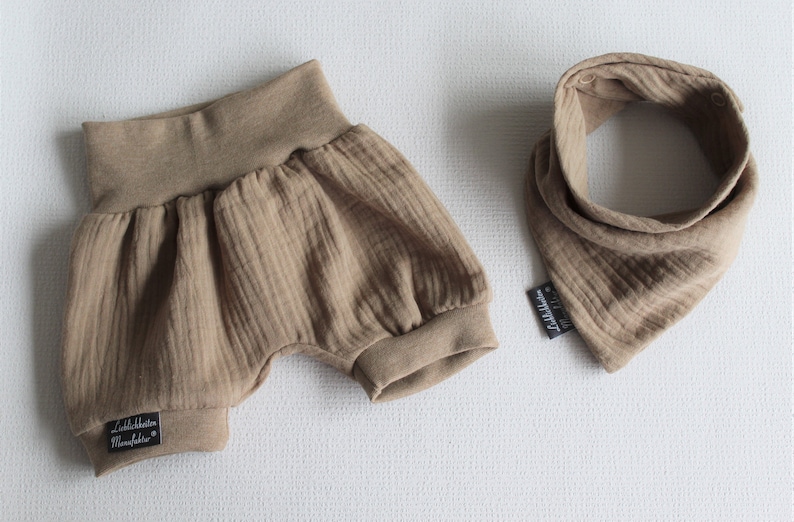May include: A brown ribbed cotton baby bib and matching shorts set. The bib has a triangular shape and the shorts have a gathered waist. Both items have a black label with the text "Littleblossom Manufaktur" on them.
