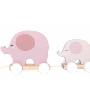 Könnte beinhalten: Zwei hölzerne Elefanten-Spielzeuge zum Ziehen in Rosa- und Weißtönen. Die Elefanten haben einen hellrosa Körper mit einem dunkleren rosa Ohr und einer weißen Radbasis. Ein weißer Faden ist am vorderen Elefanten befestigt.