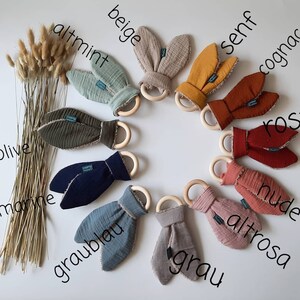 May include: Ten wooden teethers with fabric bunny ears in various colors, including beige, altmint, olive, marine, graublau, grau, altrosa, nude, ros, and cognac. The teethers are arranged in a circle on a white background.