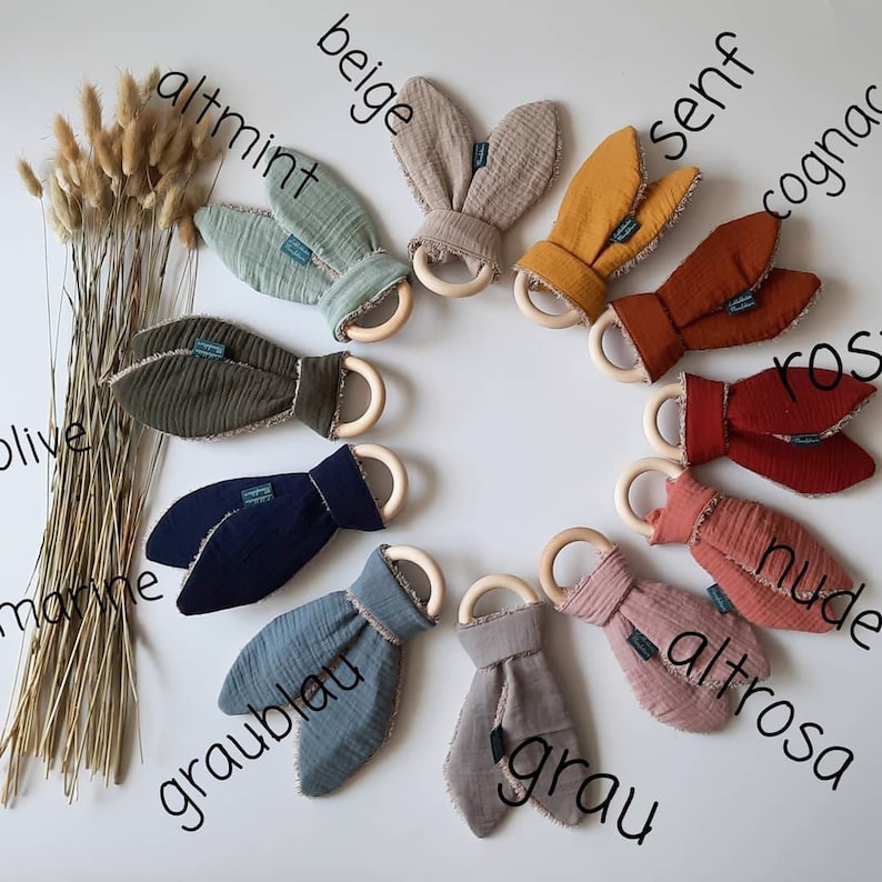 May include: Ten wooden teethers with fabric bunny ears in various colors, including beige, altmint, olive, marine, graublau, grau, altrosa, nude, ros, and cognac. The teethers are arranged in a circle on a white background.