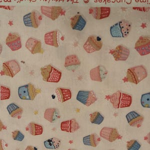 Könnte beinhalten: Weißer Stoff mit einem sich wiederholenden Muster aus bunten Cupcakes in Rosa, Blau und Braun. Die Cupcakes haben verschiedene Designs und sind über den Stoff verteilt, zusammen mit kleinen Sternen.
