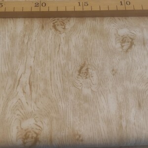 Könnte beinhalten: Nahaufnahme eines Stoffes mit Holzmaserung in Beige- und Brauntönen. Der Stoff hat eine realistische Holzstruktur mit Knoten und Maserungslinien. Ein Holzlineal oben zeigt die Maße in Zentimetern an.