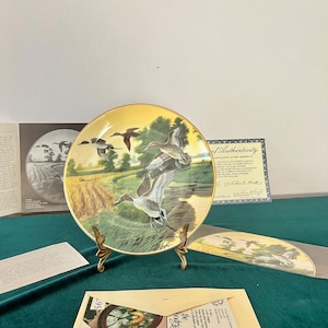 Peut inclure: Une assiette en céramique décorative avec un bord doré présente une peinture de quatre canards en vol au-dessus d'un champ vert et d'un étang. L'assiette est sur un support en métal doré. Le texte "The Heritage" est imprimé sur l'assiette.