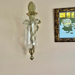 May include: A gold wall sconce with a clear glass candle holder. The sconce has a pineapple-shaped finial and a decorative design.
