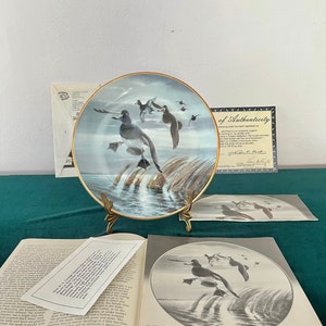 May include: A decorative ceramic plate with a gold trim featuring a painting of a blue-winged teal duck in flight over a body of water with reeds. The plate is on a gold stand and has a certificate of authenticity.