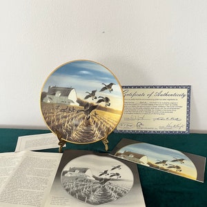 Pode incluir: Um prato de cerâmica com uma pintura de um celeiro branco num campo com gansos voando por cima. O prato está num suporte dourado e tem um certificado de autenticidade. O certificado é da Ducks Unlimited, Inc. e é assinado pelo artista, George Cutts. O texto do certificado diz: "Isto certifica que o prato que acompanha foi registado como #____ e que é um original autêntico na segunda liquidação da coleção de pratos da Ducks Unlimited, Inc. A primeira coleção de pratos alguma vez emitida com o apoio e patrocínio completos da Ducks Unlimited, Inc. Feito em fina porcelana óssea com guarnição de ouro de 22 quilates, este prato é um tesouro para colecionadores. Foi produzido numa edição limitada a um máximo de 150 disparos."