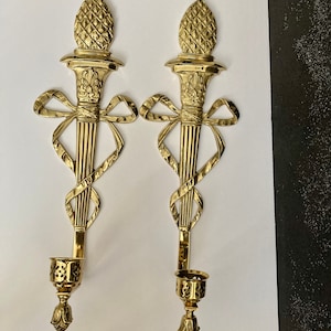 May include: A pair of gold-toned metal wall sconces with a decorative bow and a pineapple finial. Each sconce has a candle holder with a cut-out design.
