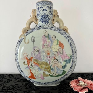 Puede incluir: Un jarrón grande, redondo, de porcelana blanca con detalles azules y dorados. El jarrón presenta una escena de ocho inmortales chinos con un tigre. El jarrón tiene dos asas en forma de dragón.