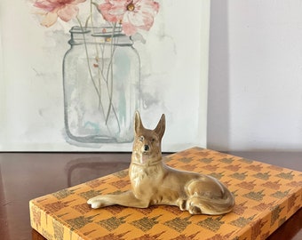 May include: A brown ceramic figurine of a German Shepherd dog lying down on a box with a repeating pattern of buildings. The box is brown with a yellow background.