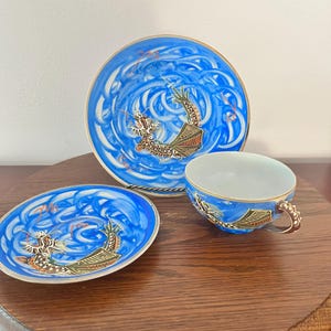 May include: A porcelain tea set consisting of a cup and two saucers, each featuring a vibrant blue swirling background with a detailed dragon illustration in black, gold, and green. The cup has a curved handle and gold-trimmed edges.