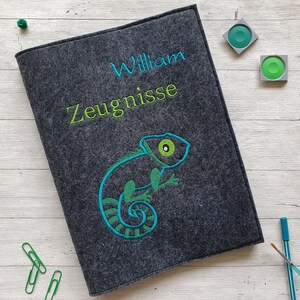 Könnte beinhalten: Ein grauer Filzordner mit dem Namen "William Zeugnisse" in grün gestickt. Auf der Vorderseite des Ordners ist ein grüner und blauer Chamäleon gestickt.