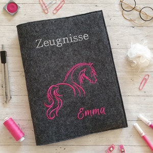 Könnte beinhalten: Ein grauer Filzordner mit dem Wort "Zeugnisse" in weiß gestickt und einer pinkfarbenen Pferdesilhouette mit dem Namen "Emma" darunter gestickt.