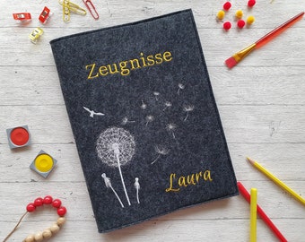 Könnte beinhalten: Ein grauer Filzordner mit weißen gestickten Löwenzahnpflanzen und dem Text "Zeugnisse" und "Laura" in gelbem Garn.