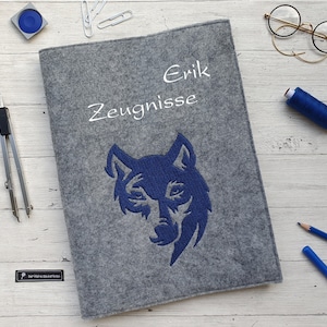Könnte beinhalten: Grauer Filzordner mit einem blauen gestickten Wolfskopf und dem Text "Erik Zeugnisse" in weiß.