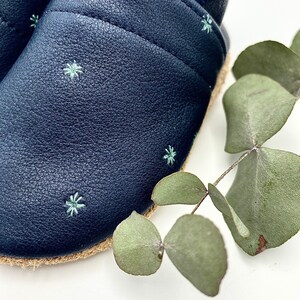 May include: A pair of dark blue leather baby shoes with a light blue star pattern. The shoes have a jute sole and are made of soft leather.