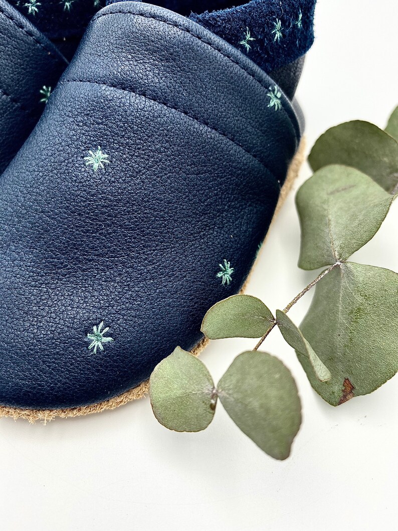 May include: A pair of dark blue leather baby shoes with a light blue star pattern. The shoes have a jute sole and are made of soft leather.