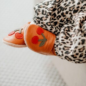 Könnte beinhalten: Orangefarbene Baby-Lederschuhe mit roten Kirschapplikationen und grünen Blättern. Die Schuhe werden an den Füßen eines Babys gezeigt, mit einer Hose mit Leopardenmuster im Hintergrund. Die Schuhe haben eine weiche, weiße Sohle.