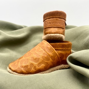 Könnte beinhalten: Ein Paar braune Leder-Babyschuhe mit einem Schaft aus Wildleder. Die Schuhe haben ein Leopardenmuster und der Schaft trägt den Text "STAY WILD" in Prägung.
