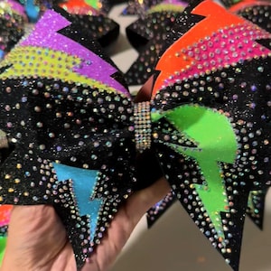 May include: A large black hair bow covered in iridescent rhinestones, featuring neon lightning bolt patterns in orange, lime green, purple, and blue. The center of the bow is cinched with a band of small, clear, sparkling crystals.